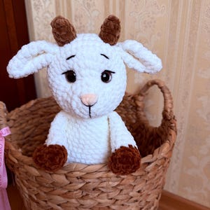 May include: A handmade white crochet goat with brown horns and hooves sits in a woven basket. The goat has large, dark eyes and a pink nose. The basket is made of natural-colored woven material.