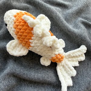 May include: A handmade plush koi carp in white and orange. The fish has a soft, textured appearance with detailed fins and tail. The koi is lying on a grey surface.