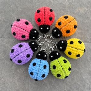 Puede incluir: Una colección de seis llaveros de mariquita tejidos a ganchillo en varios colores, incluyendo rojo, rosa, naranja, amarillo, verde, azul y morado. Cada mariquita presenta una cabeza negra con ojos blancos, un cuerpo texturizado con puntos negros y una anilla de metal.