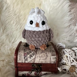 Puede incluir: Un pequeño juguete de búho de crochet con un sombrero blanco y un cuerpo marrón. El búho está sentado en una caja de madera con un diseño floral.