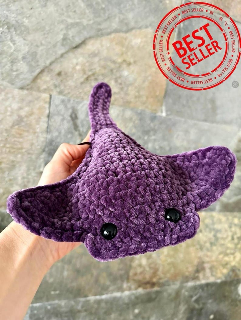 May include: Purple crocheted stingray plush toy with black eyes. The toy is being held by a hand.