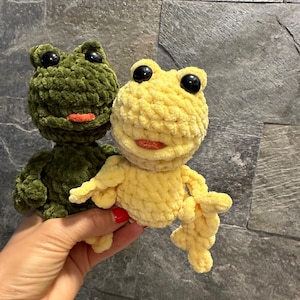 May include: Two crocheted frog plushies, one green and one yellow, with black eyes and orange mouths. The frogs are sitting on a hand.