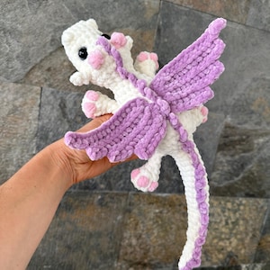 May include: A white crocheted dragon with purple wings and a long tail. The dragon has pink eyes and pink paws.