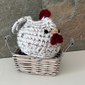 Puede incluir: Un pollo blanco de crochet con detalles rojos y amarillos se sienta en una cesta de mimbre gris.