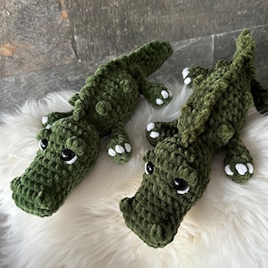 Puede incluir: Dos peluches de cocodrilo de crochet verde con garras blancas y ojos negros. Los peluches están tumbados sobre una superficie blanca y esponjosa.