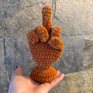 May include: A rust-colored, plush, crocheted hand sculpture featuring a raised middle finger gesture. The object is crafted from a soft, textured yarn with a visible looped stitch pattern and stands on a circular, flat base for stability.