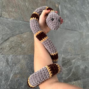 May include: A gray crocheted snake toy with brown and yellow stripes. The snake is curled up and has a cute face.