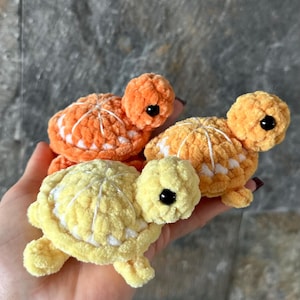 May include: Three small, plush, crocheted turtle figurines in shades of bright orange, soft yellow, and a lighter golden yellow. Each turtle features a rounded shell with white yarn detailing, a small head with a black bead eye, and four stubby legs.