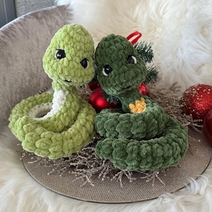 May include: Two handmade crochet snakes in shades of green. One is light green with a white underbelly, and the other is dark green. Both have black eyes and are coiled. They are placed on a decorative silver plate with holiday ornaments.