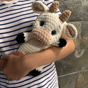 May include: A white crocheted cow plush toy with brown horns, black hooves, and pink ears. The toy is being held by a person wearing a striped shirt.