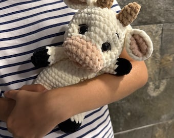 May include: A white crocheted cow plush toy with brown horns, black hooves, and pink ears. The toy is being held by a person wearing a striped shirt.