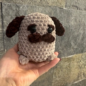 May include: A crocheted pug dog plushie in light brown with dark brown ears and mustache. The pug has large black eyes and small feet. The plushie is handmade and has a textured appearance.