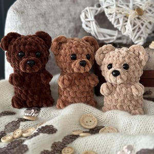 Könnte beinhalten: Drei gehäkelte Teddybären in Braun, Hellbraun und Beige. Die Bären sitzen auf einer weißen Decke mit verstreuten Holz-Knöpfen. Auf einem der Knöpfe steht der Text "Handmade with Love".