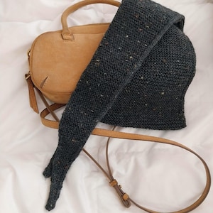 May include: A tan leather crossbody bag with a zipper closure and an adjustable strap, partially covered by a dark charcoal gray knit scarf featuring subtle multicolored flecks throughout the textured fabric of the accessory.