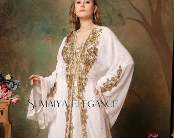 May include: A white maxi dress with gold embroidery and a tassel tie. The dress has long sleeves with bell bottoms and a V-neckline. The dress is adorned with intricate floral patterns and sequins. The text "SUMAIYA ELEGANCE" is visible on the dress.