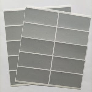 May include: Two sheets of rectangular, matte grey adhesive labels arranged in a grid pattern. Each sheet contains multiple blank, solid grey stickers with white borders, designed for labelling or organising items with a clean, uniform appearance.