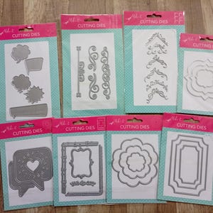 Può includere: Una collezione di fustelle in metallo argentato di varie forme, tra cui fumetti, cornici e bordi decorativi. Ogni fustella è confezionata su una carta bianca con sfondo a pois turchese e un'intestazione rosa con la scritta "CUTTING DIES".