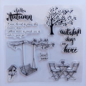 Op de afbeelding: Een set doorzichtige siliconen stempels met zwarte lijntekeningen van een boom, een vogelhuisje, een hangende schommel en een tafel met twee stoelen. De tekst bevat "hello Autumn", "autumn days are here", "Secret garden" en een kort gedicht.