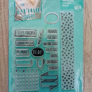 Peut inclure: Un kit de création de cartes pour les amateurs de journaux. Le kit comprend des tampons transparents avec des phrases comme "Sleep Tracker" et les jours de la semaine, un dossier de gaufrage et une matrice métallique. L'emballage est turquoise et blanc.