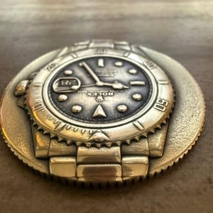 May include: A silver-coloured metal watch face with intricate detailing, including a date display at the 3 o'clock position. The watch face is set on a circular base with a textured edge, showing the watch's band and bezel.