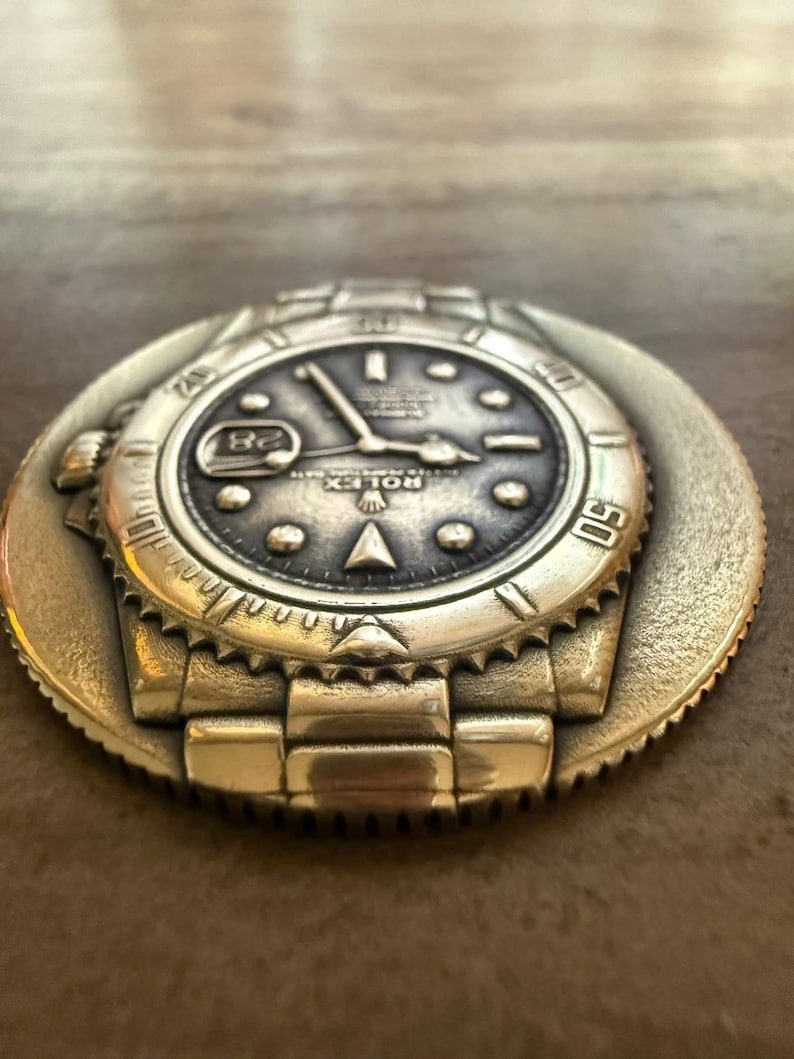 May include: A silver-coloured metal watch face with intricate detailing, including a date display at the 3 o'clock position. The watch face is set on a circular base with a textured edge, showing the watch's band and bezel.
