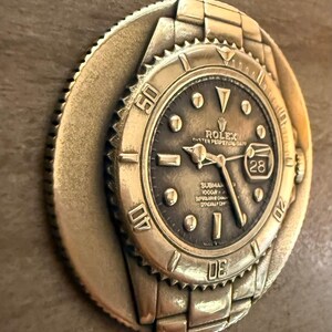 May include: A gold-coloured, circular relief sculpture of a Rolex Submariner watch. The watch face is dark with gold-coloured hands and markers. The bezel features the numbers 10, 20, 30, 40, and 50. The date is visible at the 3 o'clock position.