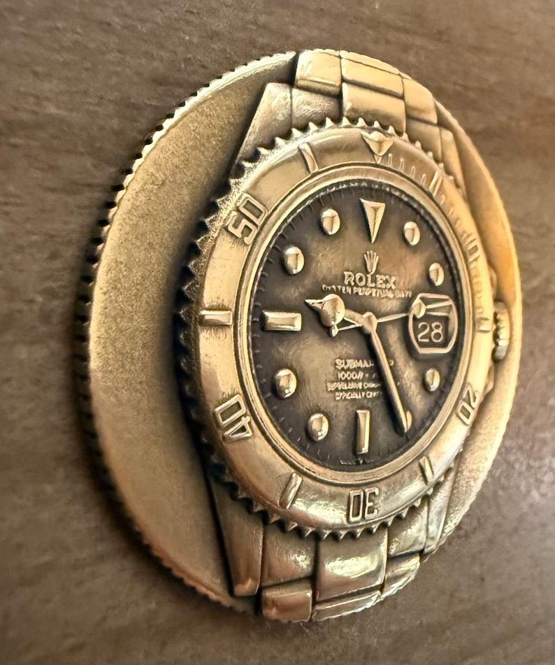 May include: A gold-coloured, circular relief sculpture of a Rolex Submariner watch. The watch face is dark with gold-coloured hands and markers. The bezel features the numbers 10, 20, 30, 40, and 50. The date is visible at the 3 o'clock position.