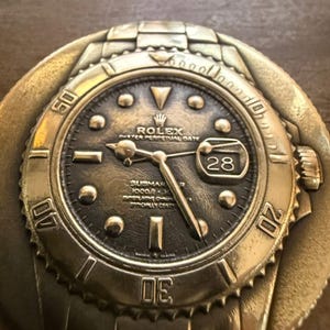 May include: A bronze-coloured metal belt buckle shaped like a Rolex Submariner watch. The watch face is dark with luminous markers, hands, and a date window at the 3 o'clock position. The bezel and band details are clearly defined.