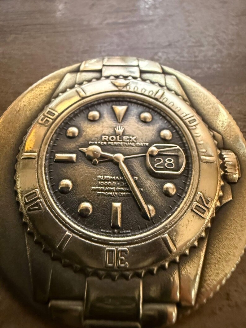 May include: A bronze-coloured metal belt buckle shaped like a Rolex Submariner watch. The watch face is dark with luminous markers, hands, and a date window at the 3 o'clock position. The bezel and band details are clearly defined.