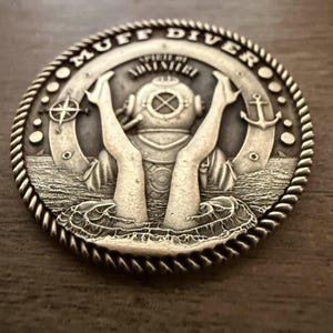 May include: A silver-colored coin featuring a vintage diving helmet and arms emerging from water. The coin's edge has a rope-like design, with the words "MUFF DIVER" and "SPIRIT OF ADVENTURE" inscribed. Nautical elements like an anchor and compass are also present.