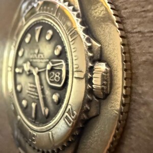 May include: A bronze-coloured relief sculpture of a Rolex watch face. The watch face shows the number 28 in a date window, with the Rolex logo visible. The bezel and crown are also visible.