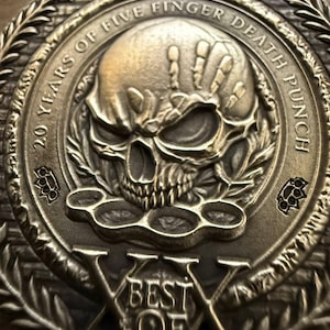 May include: A bronze-colored metal medallion with a skull and hand design. The text reads "20 YEARS OF FIVE FINGER DEATH PUNCH" and "BEST OF". The medallion features a laurel wreath border and brass knuckles.