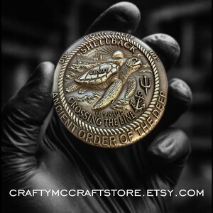 May include: A bronze-colored coin with a detailed relief design. The coin features a sea turtle, an anchor, and the text "Crossing the Line" and "Ancient Order of the Deep". The coin also has the text "Born Salty Shellback" and "US Navy".