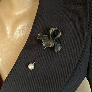 May include: A black fabric flower brooch with a pearl pin. The brooch is pinned to a black blazer.