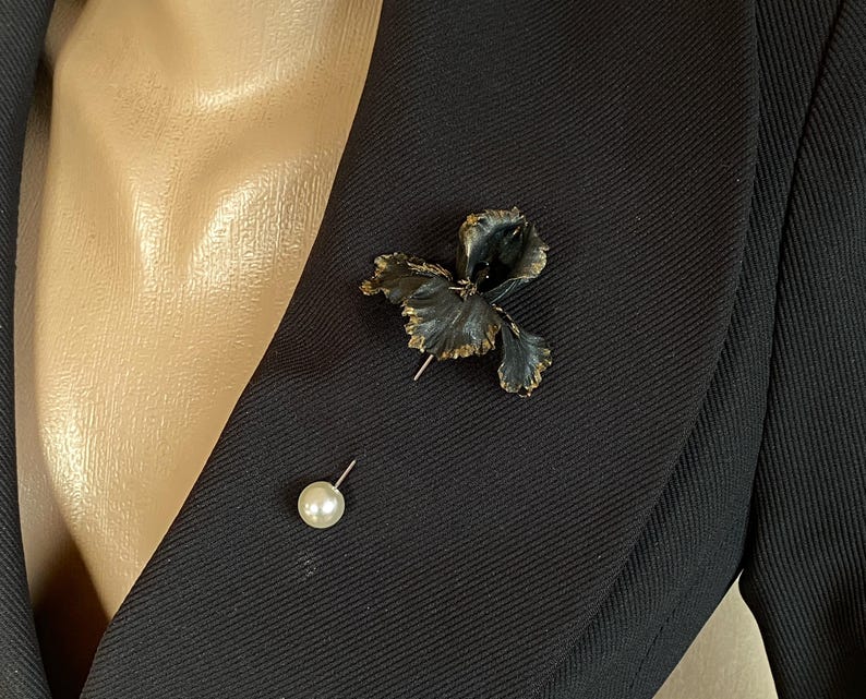 May include: A black fabric flower brooch with a pearl pin. The brooch is pinned to a black blazer.