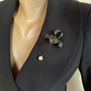 May include: A black blazer with a black and gold flower brooch and a pearl pin.