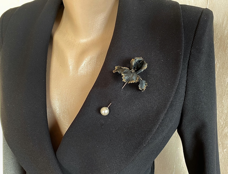 May include: A black blazer with a black and gold flower brooch and a pearl pin.
