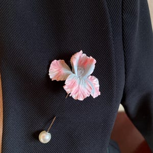 Puede incluir: Un broche de flor rosa y blanca con un acento de perla. El broche está prendido a una chaqueta negra.