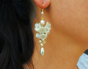 May include: White flower and pearl dangle earrings with a gold hook. The earrings are hanging from a woman's ear.