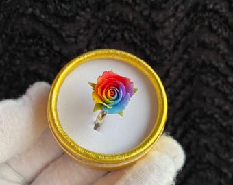 May include: A ring featuring a multi-colored rose bloom with petals transitioning from red to orange, yellow, green, blue, and purple. The ring is set into a white foam insert within a small, round, gold-colored container with a metallic finish.