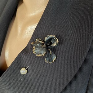 May include: A black fabric flower brooch with gold accents pinned to a black blazer. A small pearl pin is also visible on the blazer.