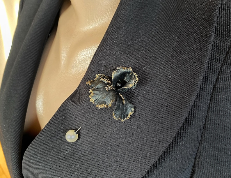 May include: A black fabric flower brooch with gold accents pinned to a black blazer. A small pearl pin is also visible on the blazer.