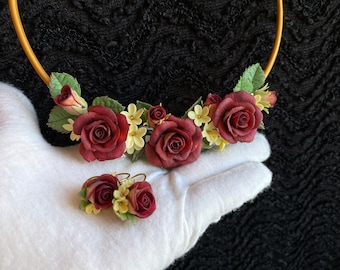 May include: A burgundy floral necklace and earrings set with small white flowers. The necklace is made of a gold hoop with burgundy roses and white flowers.