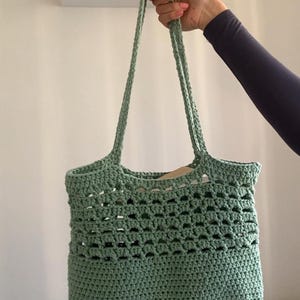 May include: A green crocheted tote bag with a wide, open top and two long straps. The bag has a delicate, open-weave pattern.