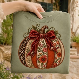 May include: A sage green crewneck sweatshirt featuring a large central graphic of a pumpkin. The pumpkin has segments decorated with leopard print, floral patterns, and solid orange, topped with a large, dark red velvet-style bow.