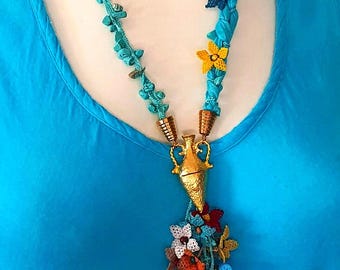 May include: A turquoise and gold necklace with a tassel of colorful crocheted flowers. The necklace features a gold pendant in the shape of a vase.