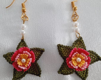 May include: A pair of dangle earrings featuring a dark olive green crocheted star base. Centered on each star is a multi-layered flower in shades of pink, yellow, and white, with a small white pearl bead at the center. The earrings hang from gold-toned hooks with two small white pearl beads on the wire.