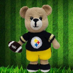 Könnte beinhalten: Ein gehäkelter Teddybär, der ein schwarz-gelbes Trikot mit einem Fußball in der Hand trägt. Das Trikot hat einen Stern und ein Fußball-Logo auf der Vorderseite.