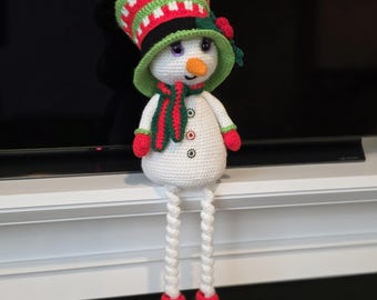 May include: A crocheted snowman doll with a festive hat, scarf, and mittens. The snowman has a white body, an orange carrot nose, and button details. The hat is red, green, and black. The snowman's legs dangle, and it is wearing green and red boots.