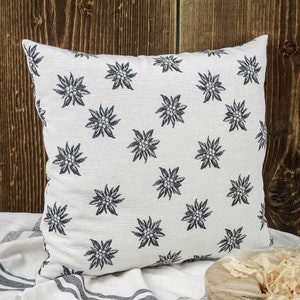 May include: White cushion with a black embroidered pattern of edelweiss flowers. The cushion is on a white and black striped fabric.